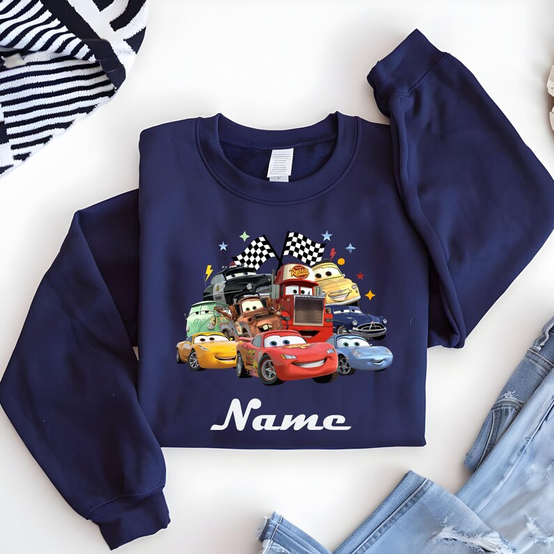 May include: Navy blue sweatshirt with a vibrant graphic of cartoon cars, including Lightning McQueen. The design incorporates checkered flags and stars. The word "Name" is written in white script below the cars. The sweatshirt is made of a soft material.