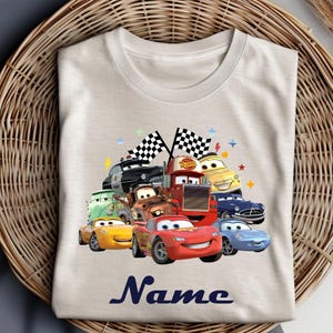 May include: A cream-coloured t-shirt with a vibrant graphic of cartoon cars, including a red race car, a tow truck, and a police car. The word "Name" is printed in a dark blue script font at the bottom of the shirt.