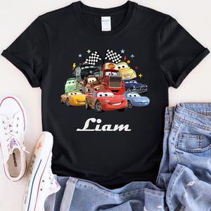 May include: Black t-shirt with a colourful graphic of animated cars, including Lightning McQueen. The name "Liam" is printed in white script below the cars. The design includes checkered flags and stars.