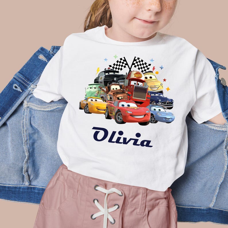May include: White t-shirt with a vibrant graphic of Cars characters, including Lightning McQueen. The name "Olivia" is printed in navy blue script below the graphic. Two checkered flags are at the top.