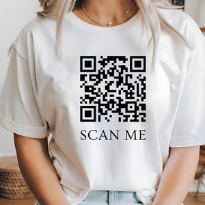 May include: White t-shirt with a large black QR code graphic and the words "SCAN ME" printed below. The shirt is a casual style, likely made of cotton or a similar fabric.