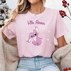 Puede incluir: Camiseta rosa con un personaje de dibujos animados de Stitch y el texto "His Angel" impreso en la parte delantera.