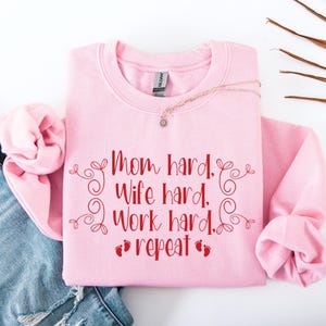 May include: A light pink sweatshirt with the text "Mom hard. Wife hard. Work hard. repeat" in red script. The design includes decorative swirls and baby footprints. A gold necklace with a pendant rests on the sweatshirt.
