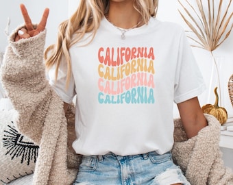 May include: White t-shirt with a graphic design of the word "CALIFORNIA" repeated four times in a rainbow color scheme. The text is in a retro, wavy font.