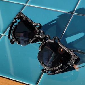 May include: Black sunglasses with a unique, ornate frame design. The sunglasses have dark lenses and are set against a blue tiled surface. The frame features a textured, swirling pattern.