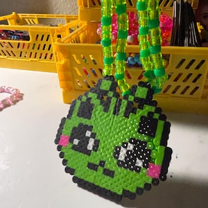 May include: A green beaded necklace with a pixelated alien pendant. The alien has black eyes, a black mouth, and pink cheeks. The necklace is made of green and clear beads. Yellow storage crates are in the background.