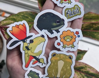 May include: A collection of colorful frog stickers in various designs. Some stickers feature frogs with flower-like features, while others depict frogs in different poses. One sticker has the word "Ribbit".