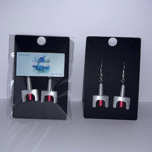 May include: Two pairs of silver-toned earrings shaped like rocket ships with red cylindrical centers. One pair is packaged in a clear plastic sleeve on a black display card with a label reading PRINT PRINCESS, while the other is on a black card.