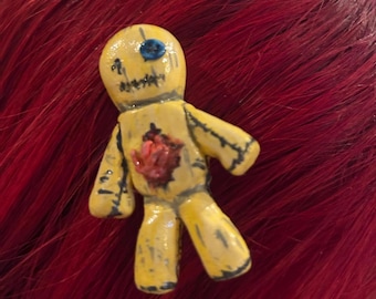 Op de afbeelding: Een gele popvormige speld met een glanzende afwerking, voorzien van één blauw oog, een gestikte mond en een rood hart met textuur op de borst. De figuur heeft zwarte, geschilderde stiknaden langs de ledematen en het hoofd voor een versleten look.