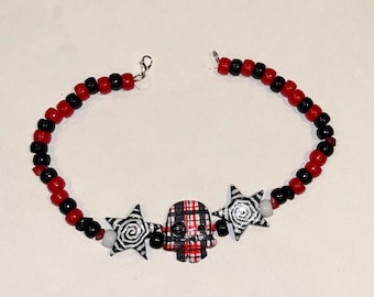 Op de afbeelding: Een kralenketting met afwisselend rode en zwarte ronde kralen, in het midden een rood-zwart geruite schedelbedel geflankeerd door twee zwart-witte stervormige bedels met spiraalpatronen en een zilverkleurige karabijnsluiting.