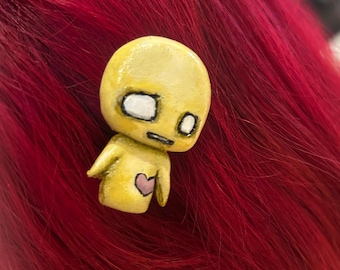 Op de afbeelding: Een klein geel figuurtje met een ronde kop, grote witte ogen met zwarte pupillen en een kleine mond. Het figuurtje heeft een roze hartje op de borst geschilderd en is geplaatst tegen een achtergrond van felrood haar.