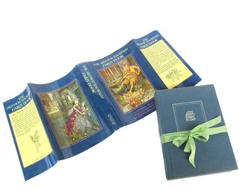 Può includere: Un libro rilegato in blu con il titolo "The Arthur Rackham Fairy Book" con illustrazioni di fate e una scena di bosco. Il libro è aperto per rivelare le pagine interne ed è legato con un nastro verde.