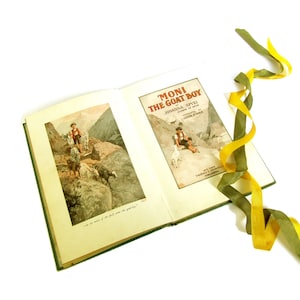 May include: An open book titled "Moni the Goat Boy" by Johanna Spyri. The book cover features a young boy with a goat in a mountainous landscape. The inside pages show a black and white illustration of a boy with goats in a mountainous landscape.