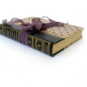 May include: A vintage hardback book with a black leather cover and gold lettering. The book is tied with a purple ribbon and has a patterned paper cover.