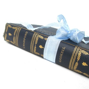May include: A navy blue hardcover book with gold lettering titled "Cranford" by "Gaskell" is tied with a light blue ribbon.