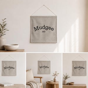 May include: A beige linen wall hanging with the word "Mudgee" and the number "2850" printed in black. The wall hanging is suspended by a natural-colored cord. The image also shows decorative vases and bowls.