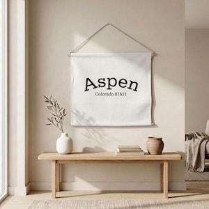 May include: A white wall hanging with the word "Aspen" in black script, with "Colorado 81611" below. The banner is suspended by a wooden dowel and a string. A wooden bench holds a vase, a small pot, and books.