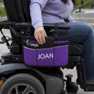 Peut inclure: Un sac latéral pour fauteuil roulant matelassé violet avec des bordures et des sangles noires, comprenant trois poches avant et un compartiment zippé. La poche centrale affiche le nom JOAN en lettres capitales blanches fixé à l'accoudoir.