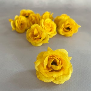 May include: A collection of bright yellow artificial rose flower heads. The fabric flowers are in various stages of bloom, with detailed petal construction. The flowers are arranged on a light gray surface, creating a visually appealing display for crafting or decorative purposes.