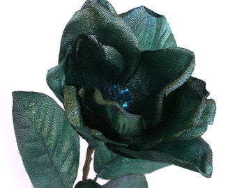May include: A single teal green artificial magnolia flower with a blue glitter center. The flower has a stem and leaves.