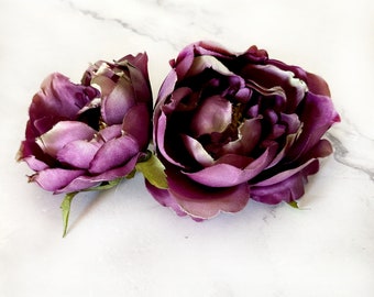 May include: Two deep purple silk peonies with a slightly ruffled texture. The peonies are on a white marble background.