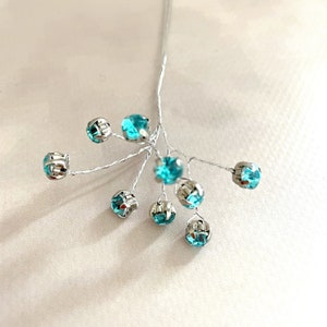 May include: A cluster of turquoise rhinestones on silver wire stems. The rhinestones are arranged in a loose, asymmetrical pattern.