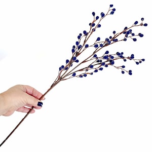 May include: A decorative floral stem featuring dark blue, fuzzy berries clustered along slender, brown branches. The stem is held against a white background, showcasing the artificial plant's details and color contrast. Ideal for adding a touch of nature to home decor.