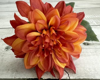 May include: A close-up of a vibrant, orange and red artificial dahlia flower. The flower has multiple layers of petals, with a gradient of colors from deep red to bright orange. A green leaf is visible on the right side of the flower. The flower is placed on a white wooden surface.
