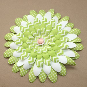 May include: A green and white fabric flower with a pink and clear rhinestone center. The flower is made of multiple layers of fabric, each with a different pattern.