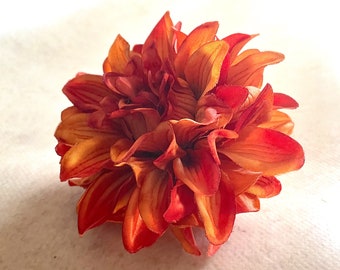 May include: A close-up of a single, artificial orange and red dahlia flower. The flower is made of fabric and has a realistic appearance.