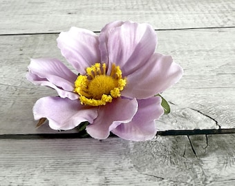 May include: A close-up of a lavender-colored artificial flower with a bright yellow center. The flower has multiple petals and green leaves at the base. The background is a light gray wooden surface.