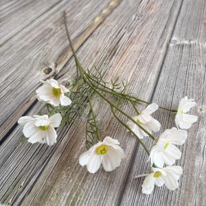 Könnte beinhalten: Ein Zweig künstlicher weißer Kosmosblumen mit gelben Zentren und grünen Stielen und Blättern. Die Blumen sind auf einer Holzoberfläche angeordnet.