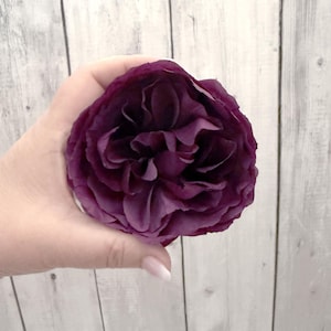 May include: A single deep purple rose with many petals, held in a hand against a white wood background.