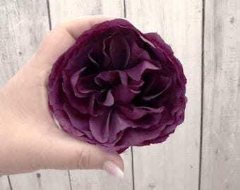 May include: A single deep purple rose with many petals, held in a hand against a white wood background.