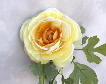 May include: A single yellow ranunculus flower with green leaves. The flower has a delicate, ruffled appearance and a soft, pastel yellow color.