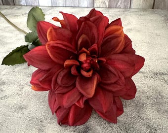 May include: A large, artificial dahlia flower in shades of deep red and orange. The flower has multiple layers of petals and a green stem with leaves. The flower is a decorative item.