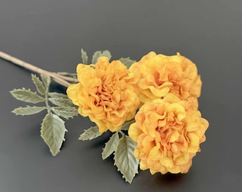 May include: A stem of three artificial marigold flowers with vibrant yellow-orange ruffled petals and serrated sage green leaves. The flowers are clustered together on a thin, light brown stem, showcasing detailed petal layers and realistic foliage.