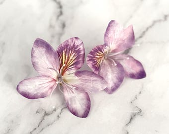 May include: Two delicate purple and white flowers with a hint of yellow in the center, arranged on a white marble surface.