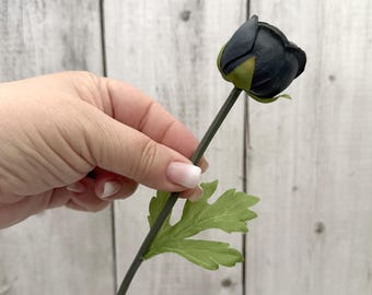 May include: A single black artificial flower with a green stem and leaf. The flower is closed and has a dark green center.