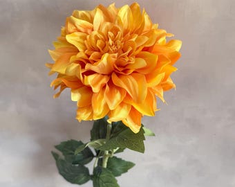 May include: A single, bright orange artificial dahlia flower with green leaves on a stem. The flower is fully bloomed and has a delicate, layered appearance.