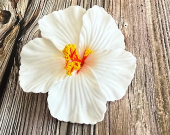 May include: A white hibiscus flower with yellow stamens and a red pistil. The flower has five large, textured petals and is set against a wooden background. The flower is a tropical floral decoration.