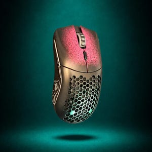 May include: A wireless computer mouse featuring a black shell with a honeycomb cutout pattern on the rear, a pink spray-painted effect on the top buttons, a silver scroll wheel, and a white logo graphic on the side panel with internal green lighting.