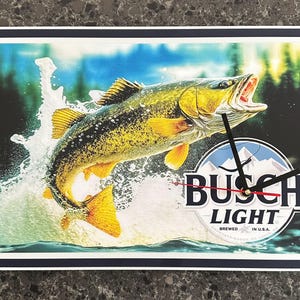 May include: Rectangular wall clock featuring a leaping fish with open mouth, water splash, and a forest background. The clock face includes the "Busch Light" logo and clock hands.