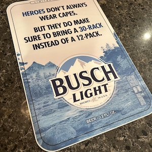 Puede incluir: Un letrero rectangular de cerveza Busch Light con una escena de montaña y agua azul y blanca. El letrero dice: "LOS HÉROES NO SIEMPRE USAN CAPAS. PERO SE ASEGURAN DE TRAER UN PAQUETE DE 30 EN LUGAR DE UNO DE 12." El letrero dice "CERVEZA 12 FL. OZ."