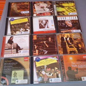 May include: A collection of 12 compact discs (CDs) with various musical artists and genres. The CDs are arranged in three rows of four. The CDs feature a variety of colors and designs, including red, blue, green, and gold. The titles of the CDs are visible, including "Season for Love", "Rouders & Hammerstein", "Morrie", "The John Tesh Project", "John Tesh", "Three Tenors", "Victoria's Secret", "Kenny G", "Andrea Bocelli", "Richard Strauss", "Carmina Burana", and "The Canticles of the Sun".