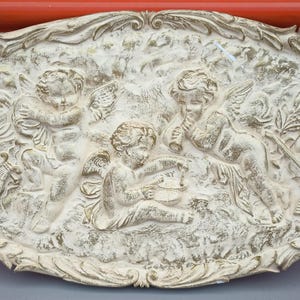 以下が含まれることがあります： 楽器を演奏するケルビムを描いた、楕円形のオフホワイトのレリーフ彫刻。詳細なアートワークには、ハープ、バイオリン、管楽器が含まれ、テクスチャード加工された背景が特徴です。ボーダーは装飾的な葉のようなデザインです。