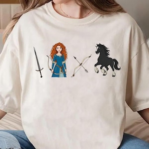 May include: Ivory-colored t-shirt featuring a cartoon illustration of a sword, a bow and arrow, a woman in a blue dress, and a black horse. The word "Ivory" is printed at the bottom of the shirt.