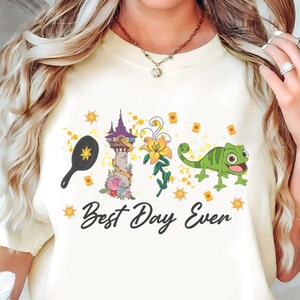 Könnte beinhalten: Ein cremefarbenes Kurzarm-T-Shirt mit einem Grafikdruck, der eine schwarze Bratpfanne, einen hohen Steinturm, eine gelbe Blume und ein grünes Chamäleon zeigt. Der Text "Best Day Ever" ist in schwarzer Schreibschrift unter den Motiven gedruckt.