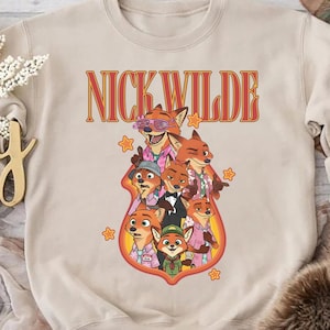 May include: Beige crewneck sweatshirt featuring a graphic of Nick Wilde and other characters from Zootopia. The text "NICK WILDE" is in orange and red lettering above the characters. The design includes stars and a cartoon style.
