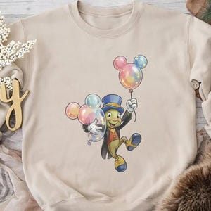 May include: Beige crewneck sweatshirt featuring a cartoon illustration of Jiminy Cricket holding Mickey Mouse-shaped balloons. The balloons are pastel colors. The character is wearing a top hat, suit, and shoes. The word "Joy" is visible in the background.